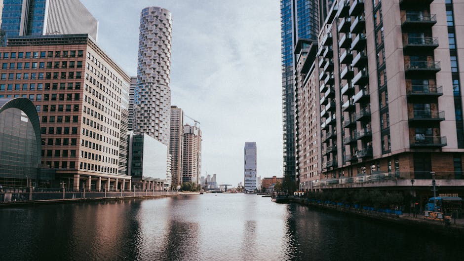 A wide view of the Canary Wharf skyline, featuring modern high-rise office buildings with glass and steel facades, situated along a calm river. The buildings exhibit a mix of architectural styles, including rectangular, cylindrical, and irregular shapes. Reflective surfaces on the structures mirror the partly cloudy sky above. The river in the foreground has a smooth, dark surface with subtle ripples, and the scene is illuminated by natural daylight, highlighting the urban environment. The image captures the clean, professional atmosphere typical of the Docklands E14 area, emphasizing the area’s contemporary architecture and waterfront setting, suitable for illustrating residential or commercial cleaning services by Carpet Cleaners E8.
