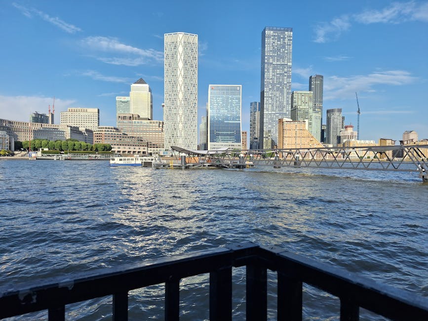 A view of the modern Canary Wharf skyline with tall glass skyscrapers overlooking the River Thames. The foreground features a black metal railing along the waterfront, with rippling water in the river. The buildings include sleek, reflective surfaces, and the sky above is blue with scattered clouds, suggesting a clear, sunny day. The image captures the urban landscape, highlighting the contemporary architecture and clean waterfront environment. This scene showcases the importance of surface cleaning and maintenance in commercial districts like Docklands E14, as emphasized by Carpet Cleaners E8 in their professional cleaning services for the area.