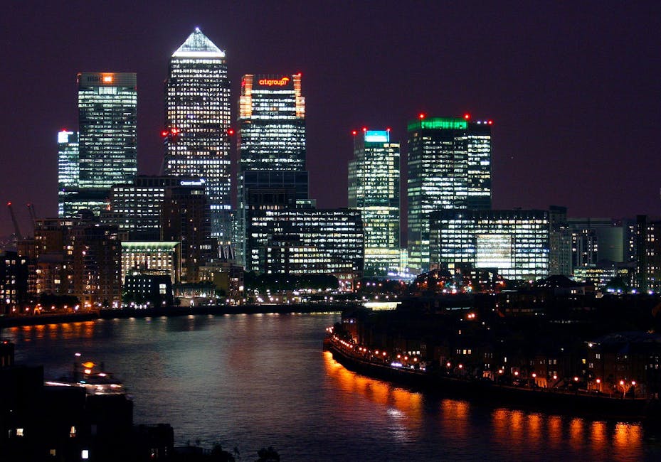 A wide view of the Canary Wharf skyline, featuring modern high-rise office buildings with glass and steel facades, situated along a calm river. The buildings exhibit a mix of architectural styles, including rectangular, cylindrical, and irregular shapes. Reflective surfaces on the structures mirror the partly cloudy sky above. The river in the foreground has a smooth, dark surface with subtle ripples, and the scene is illuminated by natural daylight, highlighting the urban environment. The image captures the clean, professional atmosphere typical of the Docklands E14 area, emphasizing the area’s contemporary architecture and waterfront setting, suitable for illustrating residential or commercial cleaning services by Carpet Cleaners E8.