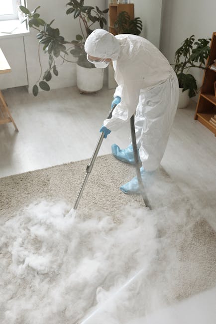 A person vacuuming an ornate, patterned area rug with a black canister vacuum cleaner in a well-lit room. The carpet features floral motifs in muted colors, and the individual's white shoes and dark t