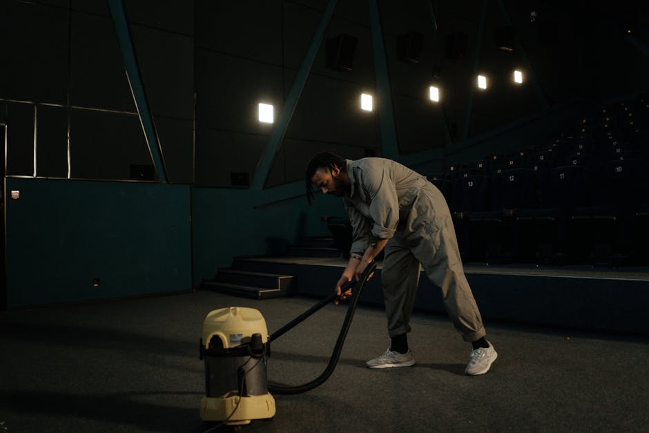 A person kneeling on a beige patterned carpet in a residential setting, preparing a portable vacuum cleaner for surface cleaning. The individual, wearing a khaki jacket and blue jeans, is opening the 