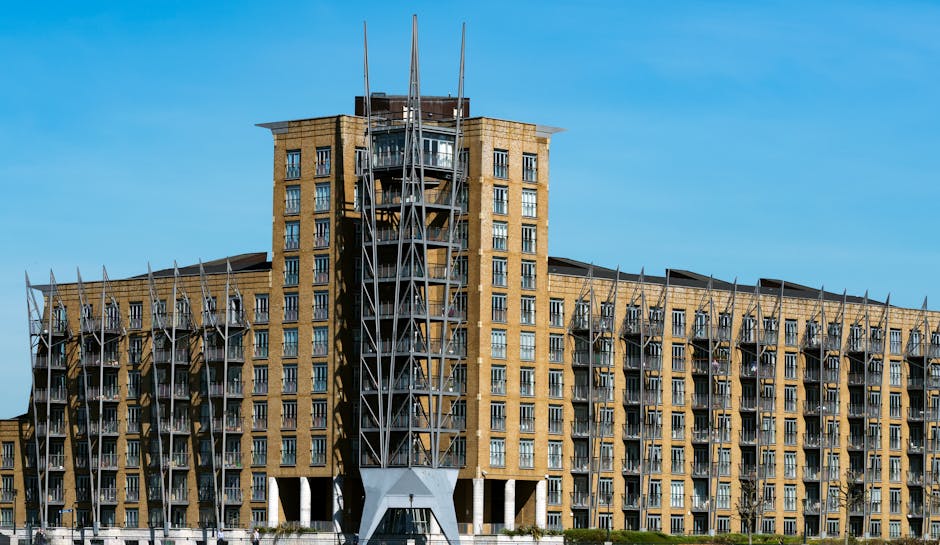 A modern residential building with multiple floors featuring yellow brick facades, glass balcony railings, and large windows, situated along a waterfront with trees in the foreground. Several boats, i